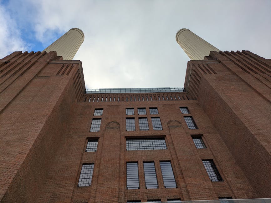 A low-angle view of the exterior of Battersea Power Station, showcasing its large brick façade with numerous rectangular multi-pane windows. The image captures two tall, cylindrical chimneys extending upward towards a partly cloudy sky. The building's brickwork includes vertical and horizontal architectural details, with some areas featuring decorative brick patterns. The scene suggests an urban environment where home relocation and furniture transport may be required, with the building serving as a reference point for local removals services. The lighting indicates daytime with natural light illuminating the structure, emphasizing the industrial architectural style common in historic power stations. This perspective illustrates the scale and grandeur of the structure, relevant to the planning of moving logistics around such iconic landmarks, as highlighted in the Battersea Power Station removals local guide by Man with Van Battersea.