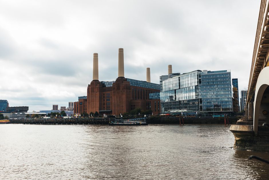 View of the historic Battersea Power Station with its distinctive four tall chimneys and red brick exterior, situated along the river Thames in London. To the right, part of a modern glass-fronted building can be seen, reflecting the cloudy sky. In the foreground, the river water appears calm, with a dock or quay area visible along the shoreline. On the right edge, a section of a vehicle, possibly a van used for house removals, is parked on the pavement near the riverbank, next to the entrance of a property. The scene is set during daytime with overcast weather, highlighting the industrial architecture of the power station and its surroundings. This image represents the kind of urban environment where professional moving services, such as those provided by Man with Van Battersea, are often involved in local home relocation and furniture transport activities, including packing, loading, and transporting belongings across the area.