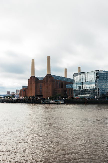 A low-angle view of the exterior of Battersea Power Station, showcasing its large brick façade with numerous rectangular multi-pane windows. The image captures two tall, cylindrical chimneys extending upward towards a partly cloudy sky. The building's brickwork includes vertical and horizontal architectural details, with some areas featuring decorative brick patterns. The scene suggests an urban environment where home relocation and furniture transport may be required, with the building serving as a reference point for local removals services. The lighting indicates daytime with natural light illuminating the structure, emphasizing the industrial architectural style common in historic power stations. This perspective illustrates the scale and grandeur of the structure, relevant to the planning of moving logistics around such iconic landmarks, as highlighted in the Battersea Power Station removals local guide by Man with Van Battersea.