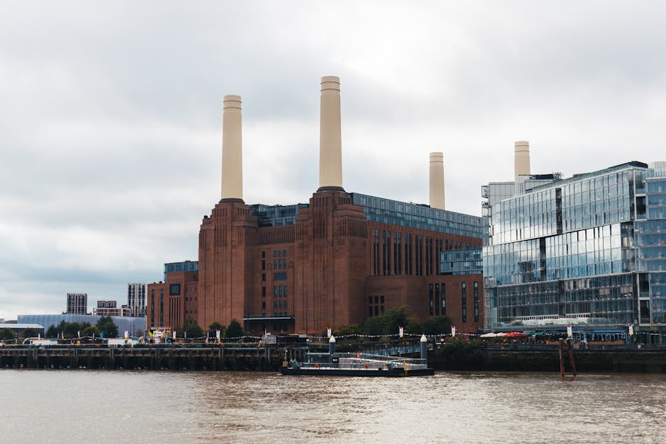 A large red-brick building with four prominent tall white chimneys situated along the riverbank in Battersea, London. The building features multiple windows and modern glass extensions on its upper le